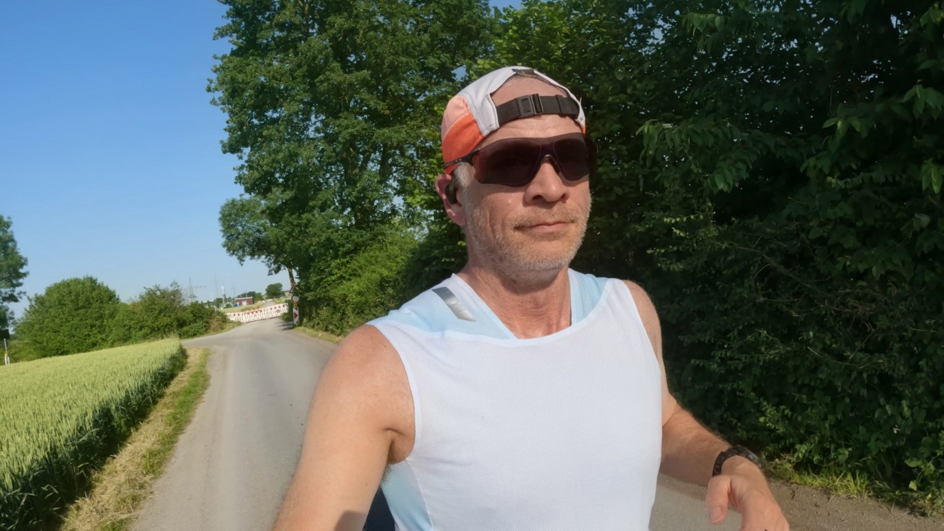 Middle-aged man running or cycling on a sunny rural road, wearing a white sleeveless shirt, orange-and-white cap, dark sunglasses, and wireless earbud, with green trees and a hedge-lined path in the background.