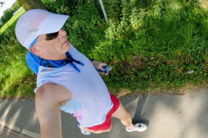 Person taking a high-angle selfie while walking on a paved path, wearing a white sleeveless shirt, red shorts, white sneakers, a light-blue neck scarf, sunglasses, and a white sun visor under green trees.