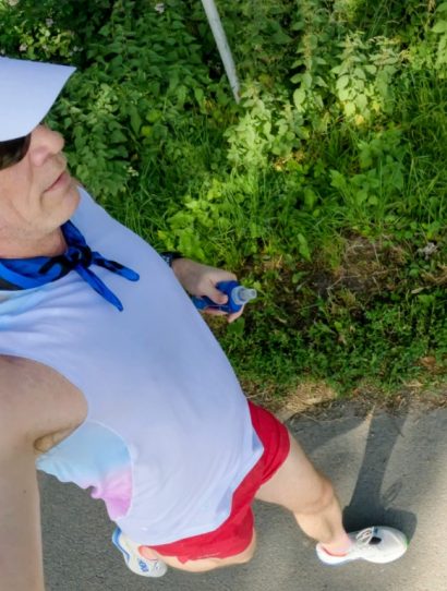 Person taking a high-angle selfie while walking on a paved path, wearing a white sleeveless shirt, red shorts, white sneakers, a light-blue neck scarf, sunglasses, and a white sun visor under green trees.