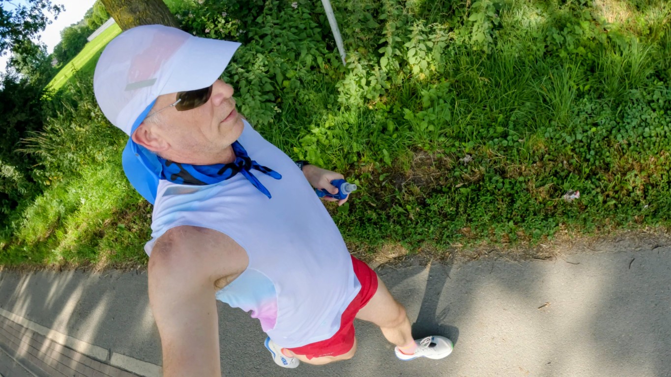 Person taking a high-angle selfie while walking on a paved path, wearing a white sleeveless shirt, red shorts, white sneakers, a light-blue neck scarf, sunglasses, and a white sun visor under green trees.