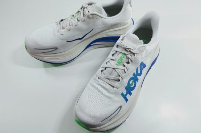 White HOKA running shoes with blue logo and green accents on a white surface, angled to show side and toe detail.