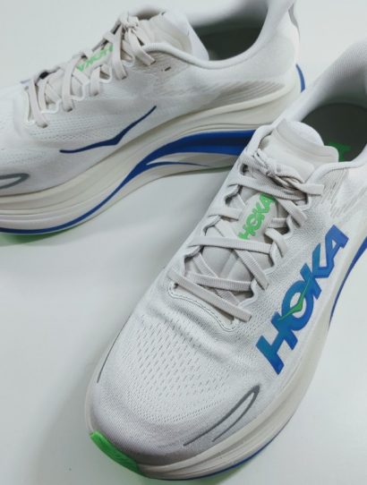 White HOKA running shoes with blue logo and green accents on a white surface, angled to show side and toe detail.