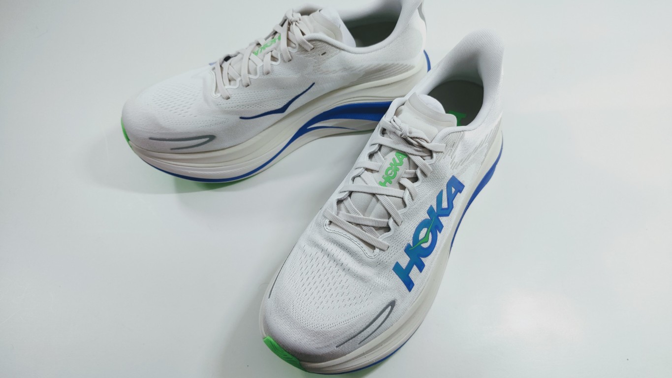 White HOKA running shoes with blue logo and green accents on a white surface, angled to show side and toe detail.
