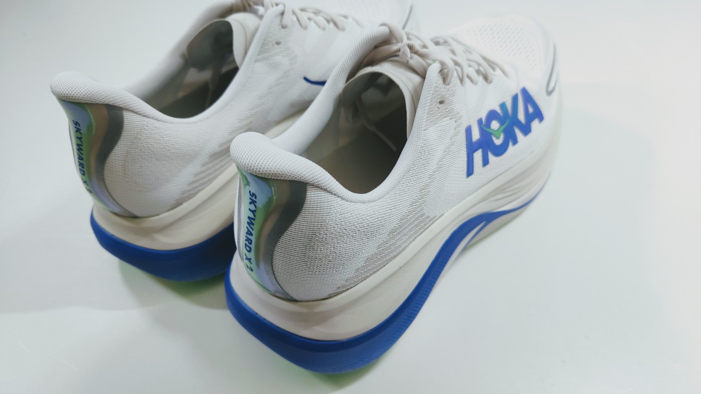 White Hoka running shoes with blue outsole, back view, showing logo on the side and heel tab