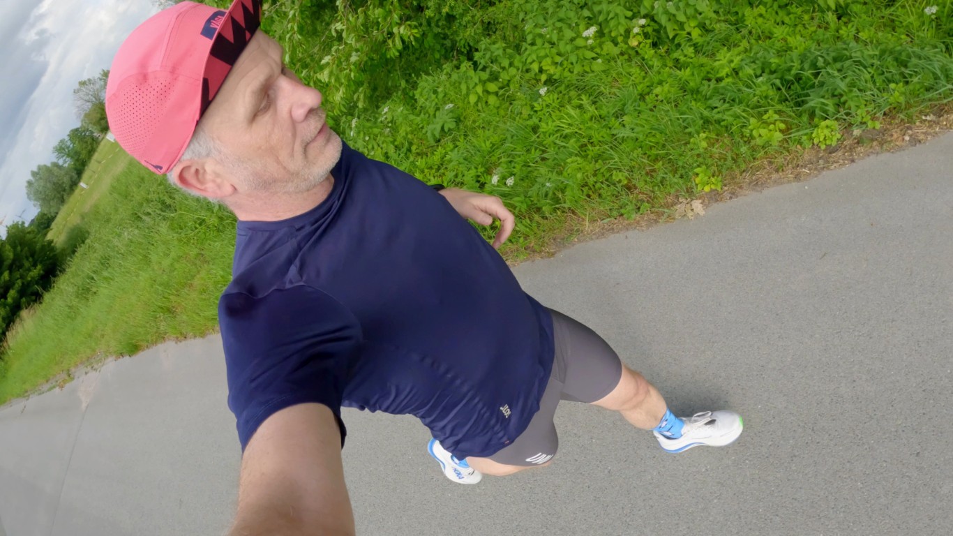 Man in a pink cap, navy shirt, and gray shorts running along a paved path with green bushes on the side, photographed from above (selfie view).