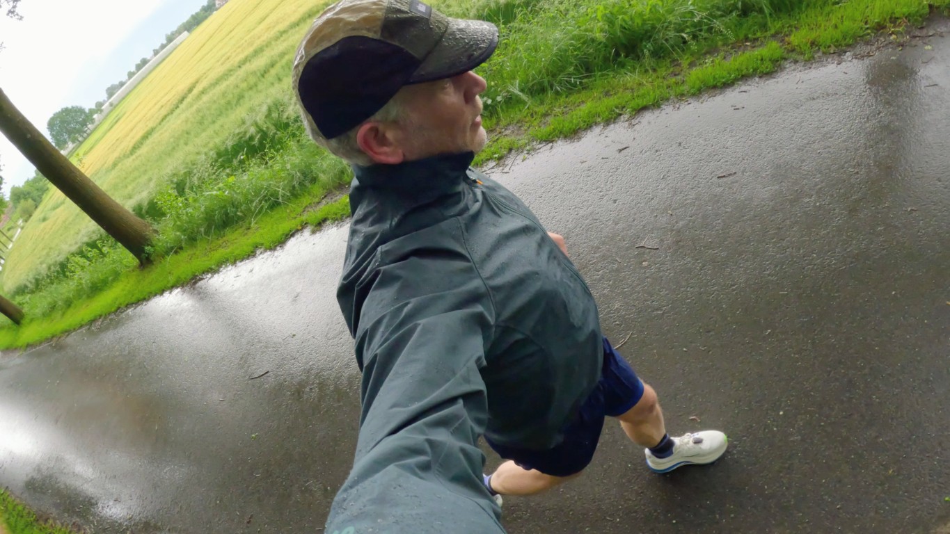 Person taking a selfie while running on a wet paved path in a green field, wearing a camo cap, gray rain jacket, navy shorts, and white sneakers.