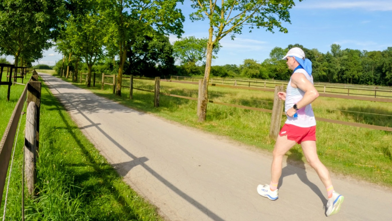 Male runner in a white tank top and red shorts jogs along a sunlit country road bordered by fences and trees.