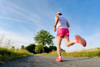Back view of a runner in a white tank top and pink shorts mid-stride on a rural path with tall grass and blue sky