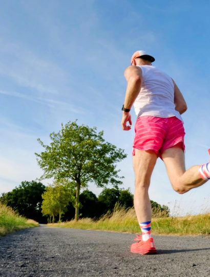 Back view of a runner in a white tank top and pink shorts mid-stride on a rural path with tall grass and blue sky