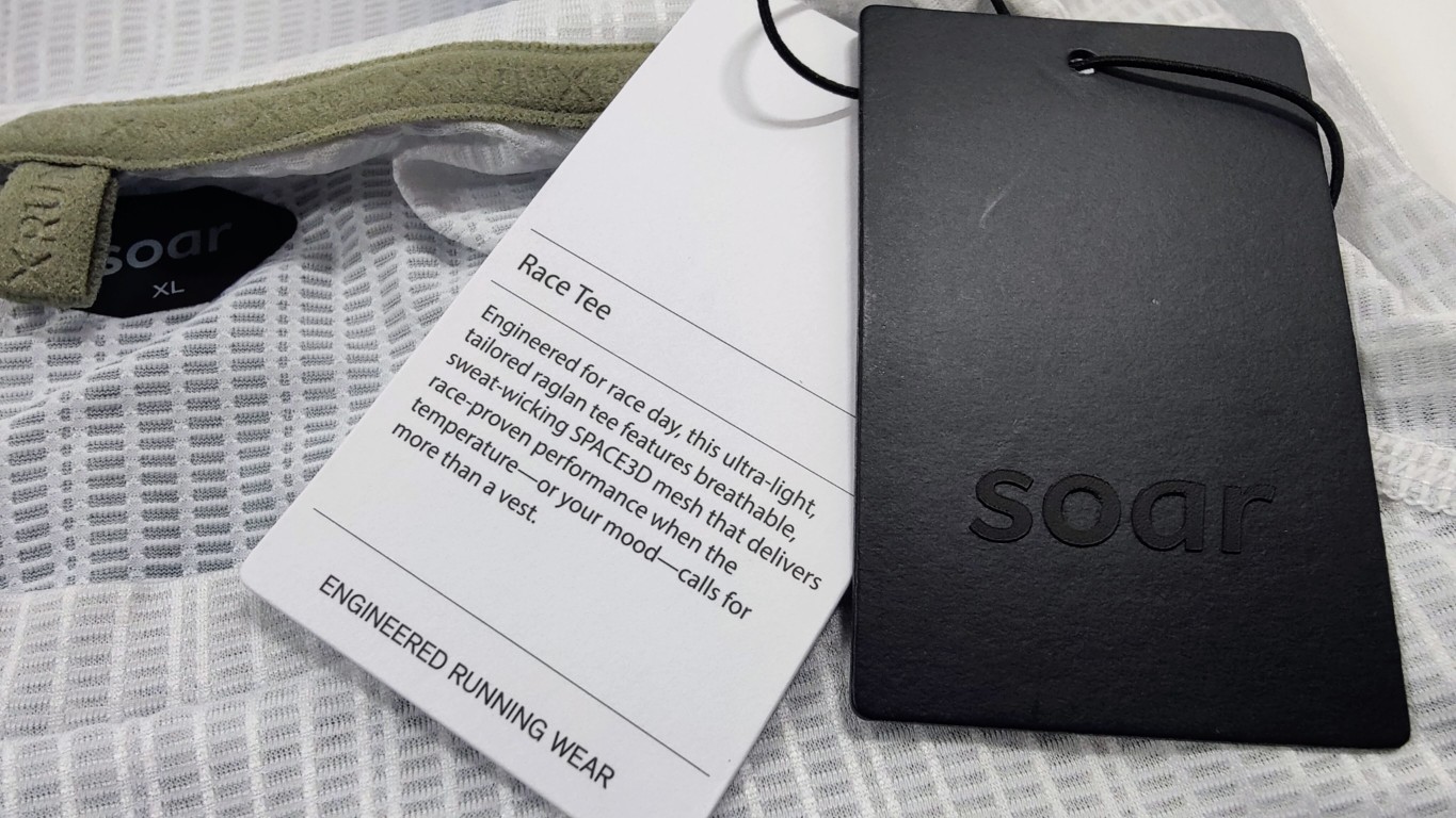 White and black hang tags reading 'Race Tee' and 'soar' lying on top of a white mesh shirt.