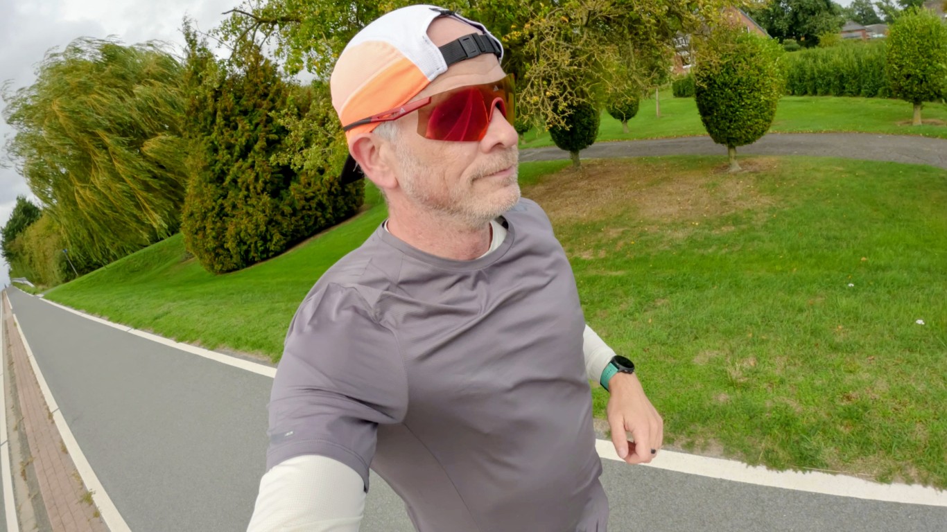 Runner taking a wide-angle selfie on a paved path, wearing a grey T-shirt over a cream long-sleeve shirt, an orange and white cap and red mirrored sunglasses.