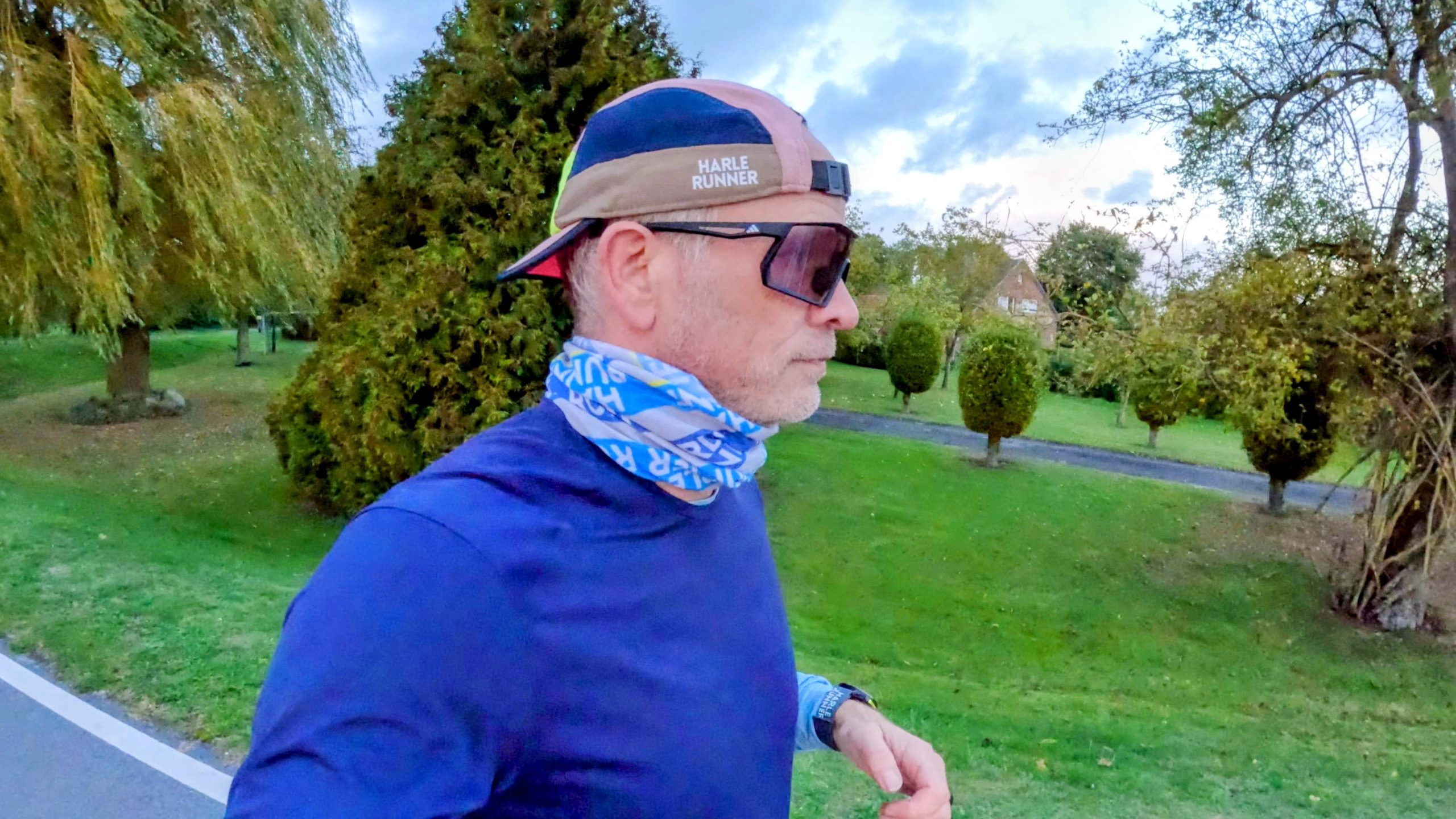 Runner in profile wearing black sports sunglasses with a violet photochromic lens, cap and neck gaiter on a road in an overcast autumn evening