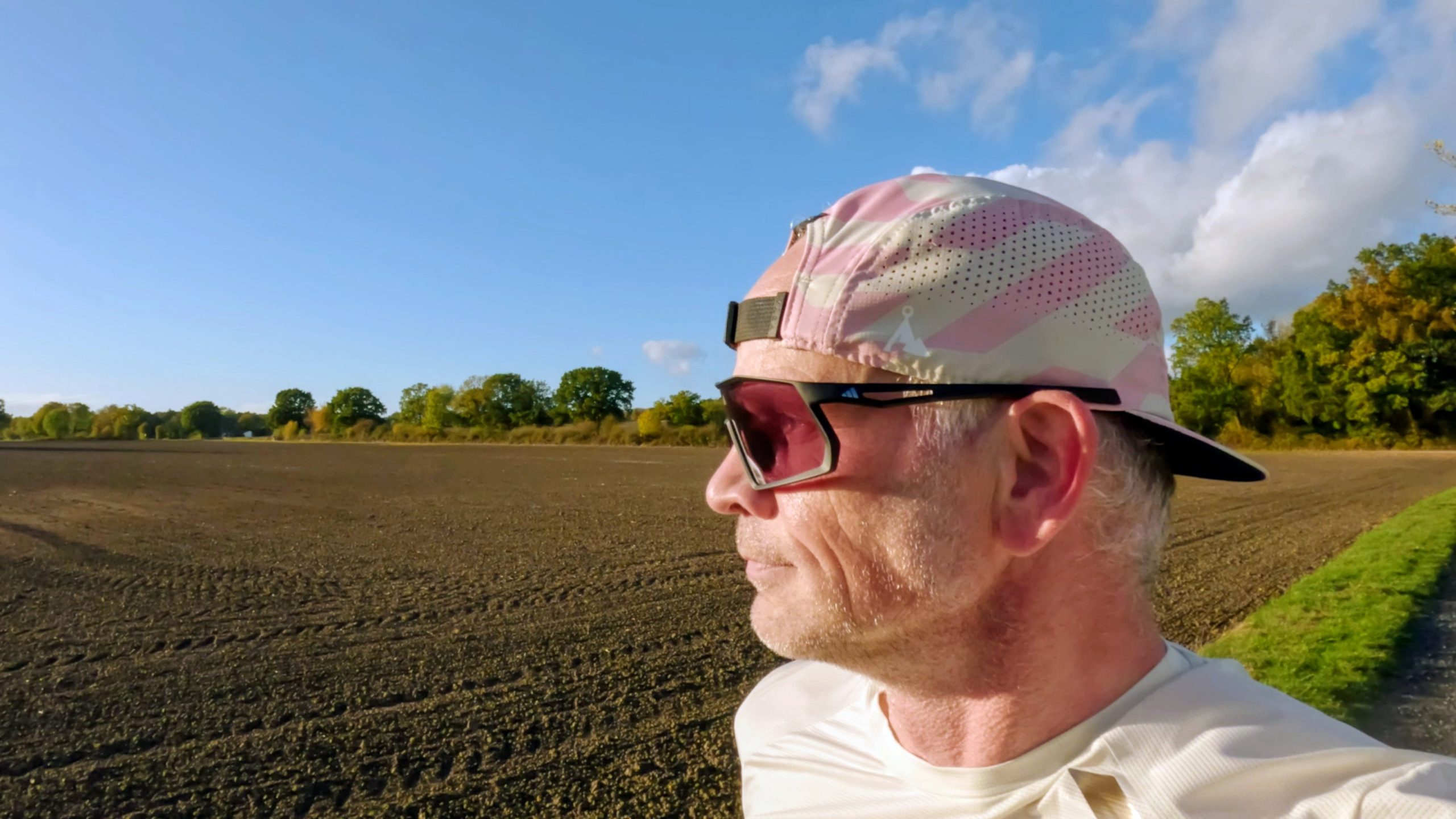 Runner in profile with black adidas Kentro sunglasses and pink-tinted lens in front of a plowed field under a blue sky