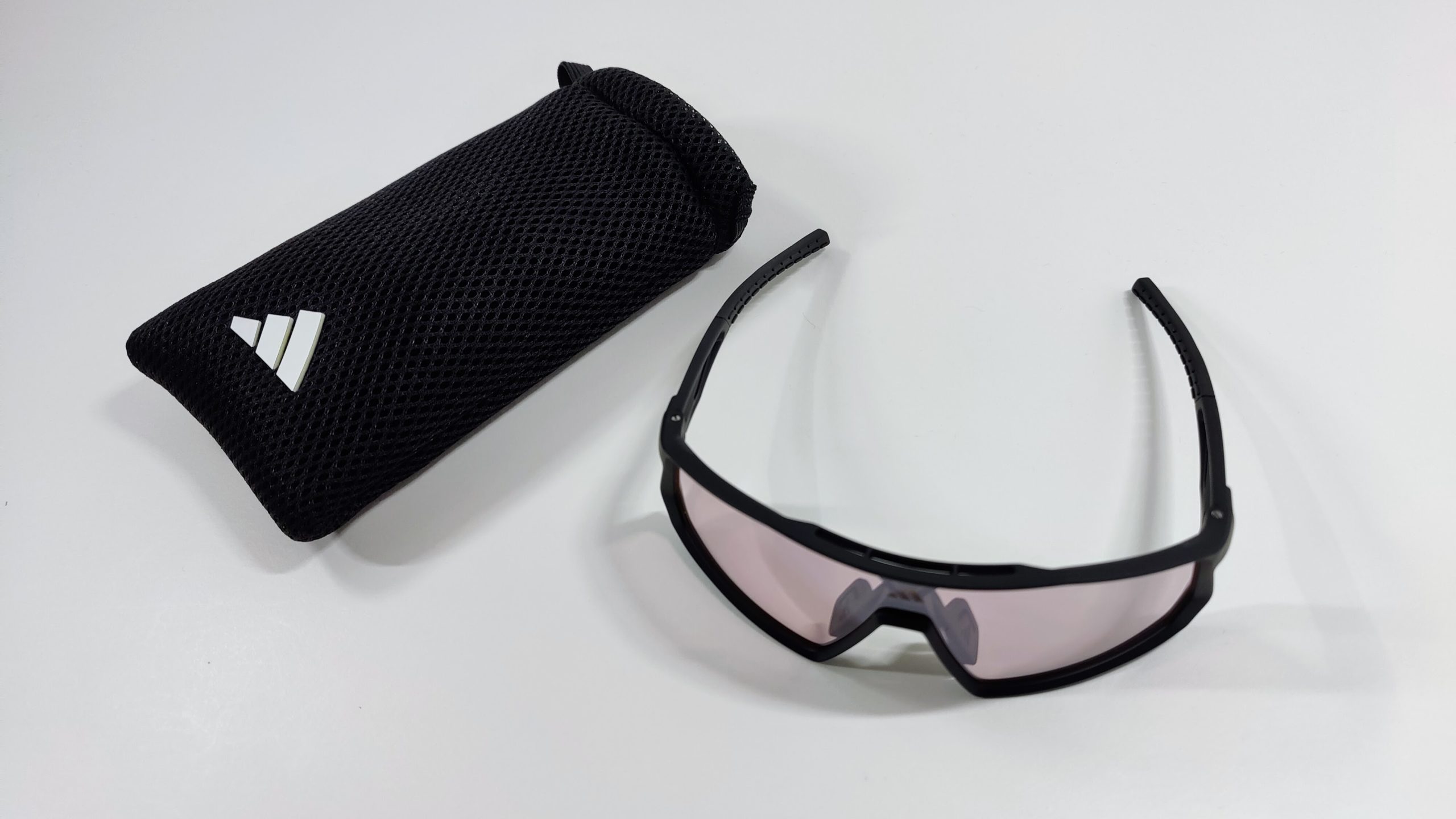 Black mesh glasses pouch with white logo next to black sports sunglasses with light violet lens on a white surface