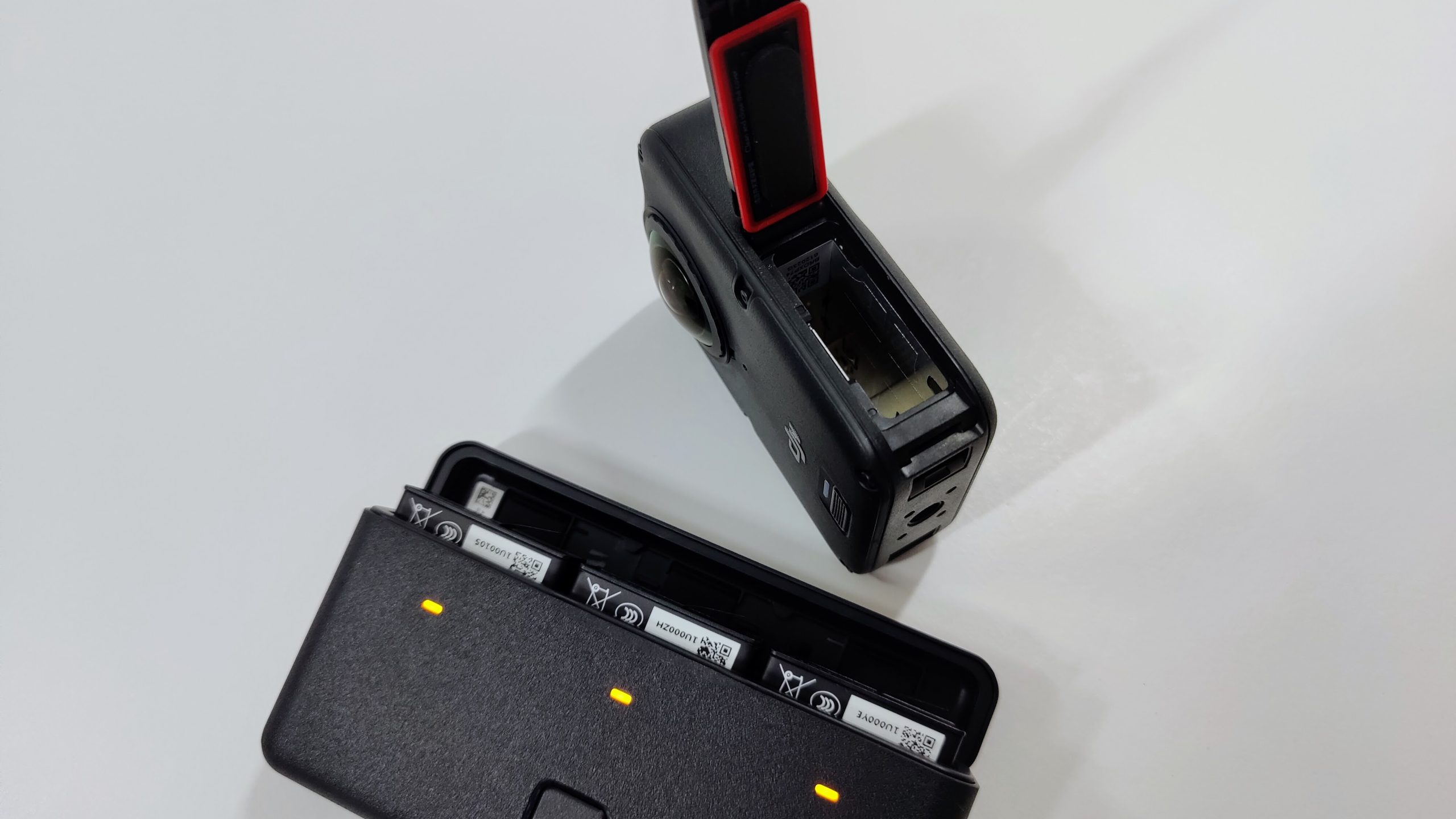 360 camera with battery compartment open next to a three-slot battery charging case with yellow LEDs.