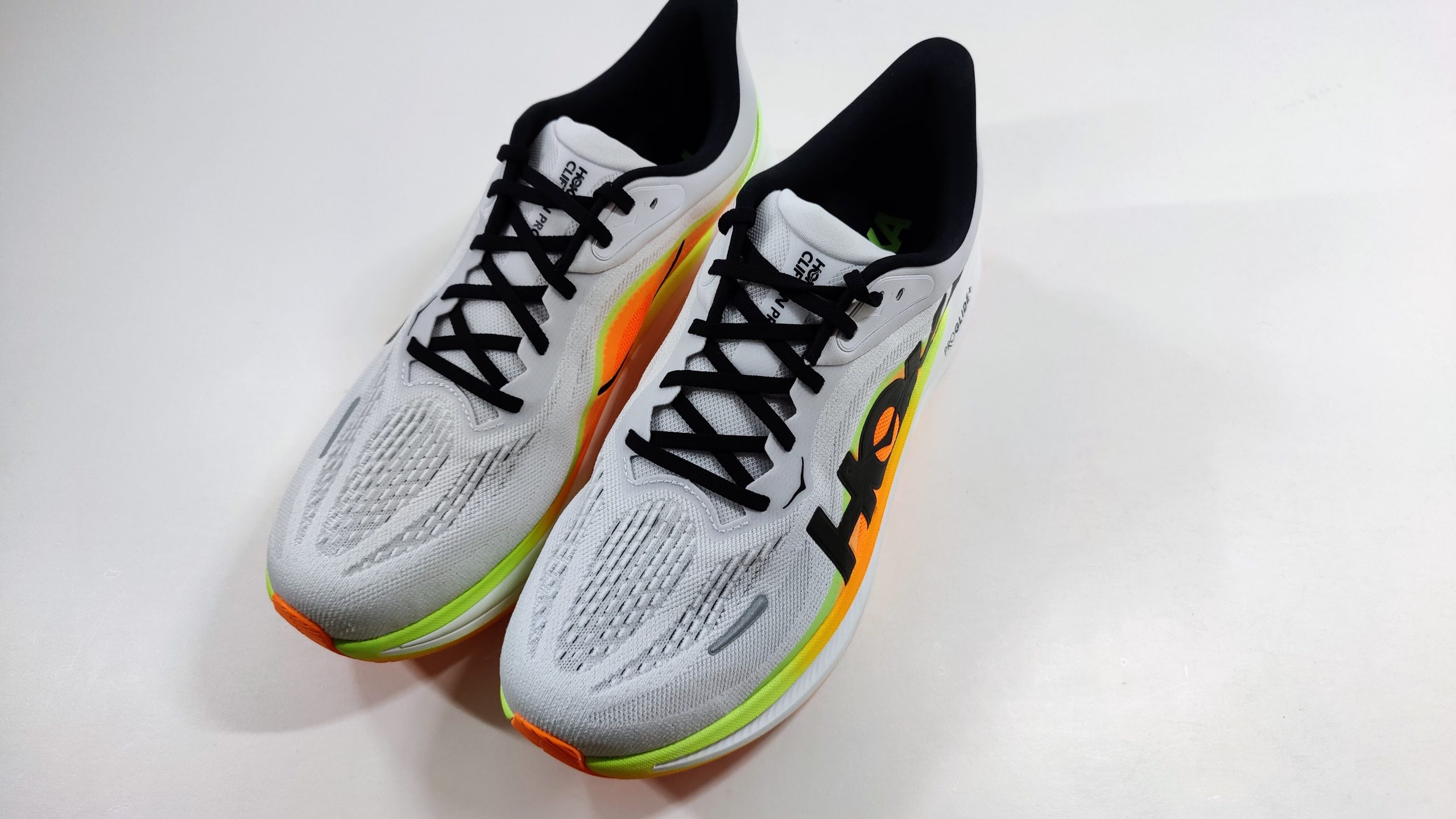 Pair of white running shoes with neon orange and lime green midsole, black laces and large black logo, photographed from above at an angle on a white surface.
