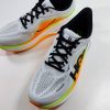 Pair of white running shoes with orange and lime green midsole seen from a higher angle, with black laces and the Hoka logo on the side.