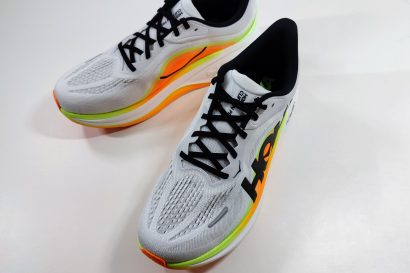 Pair of white running shoes with orange and lime green midsole seen from a higher angle, with black laces and the Hoka logo on the side.