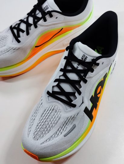 Pair of white running shoes with orange and lime green midsole seen from a higher angle, with black laces and the Hoka logo on the side.