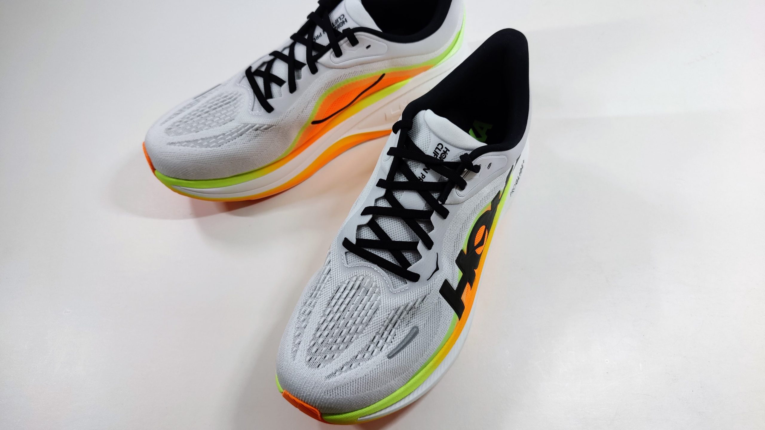 Pair of white running shoes with orange and lime green midsole seen from a higher angle, with black laces and the Hoka logo on the side.