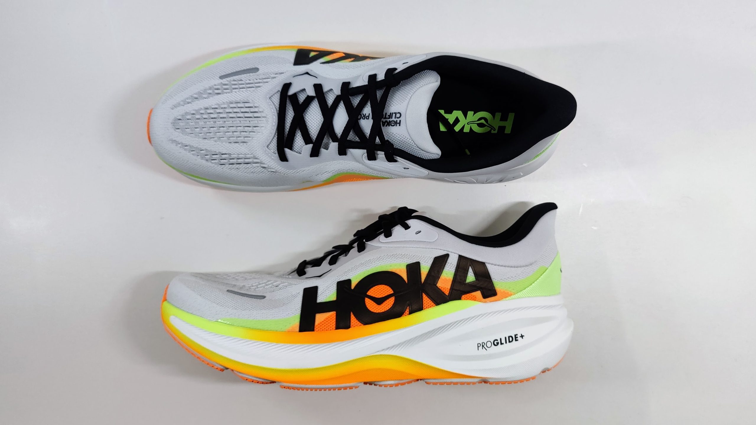 One white running shoe seen from above next to a second shoe in side profile, showing a thick neon orange and lime green midsole with a rocker shape.