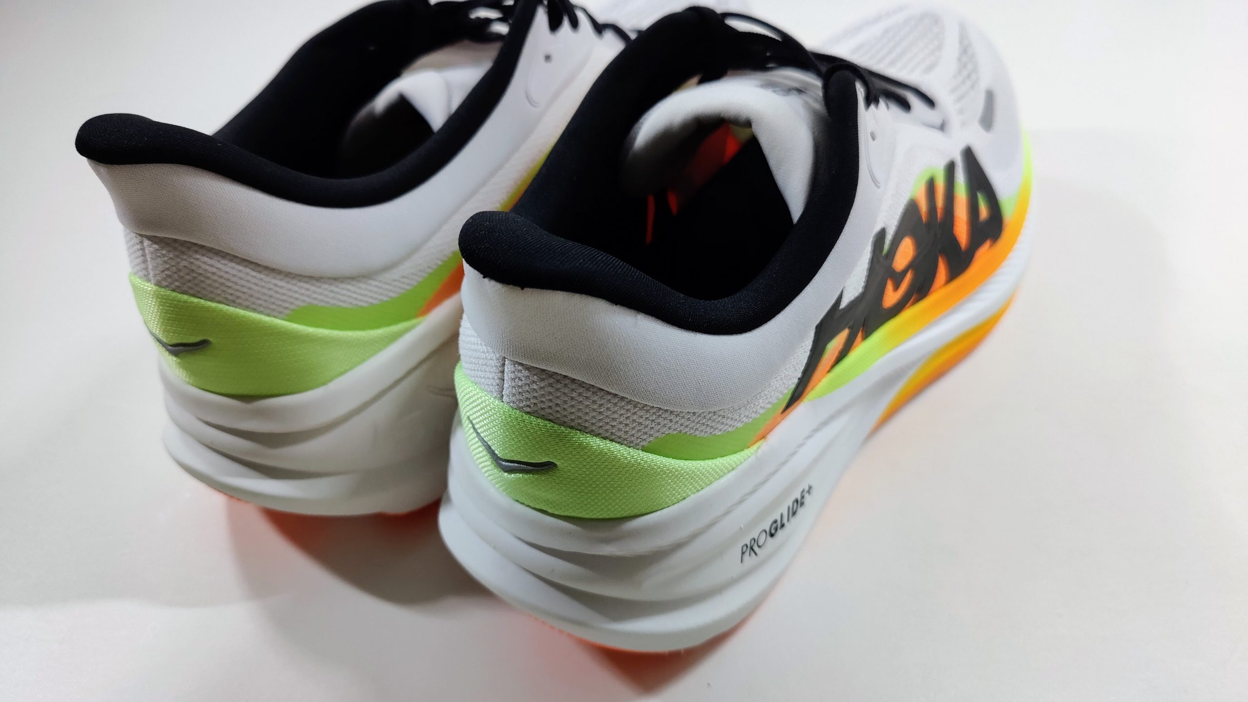Rear view of a pair of white running shoes with black padded collar, lime green heel overlay and thick white heel midsole, on a white surface.