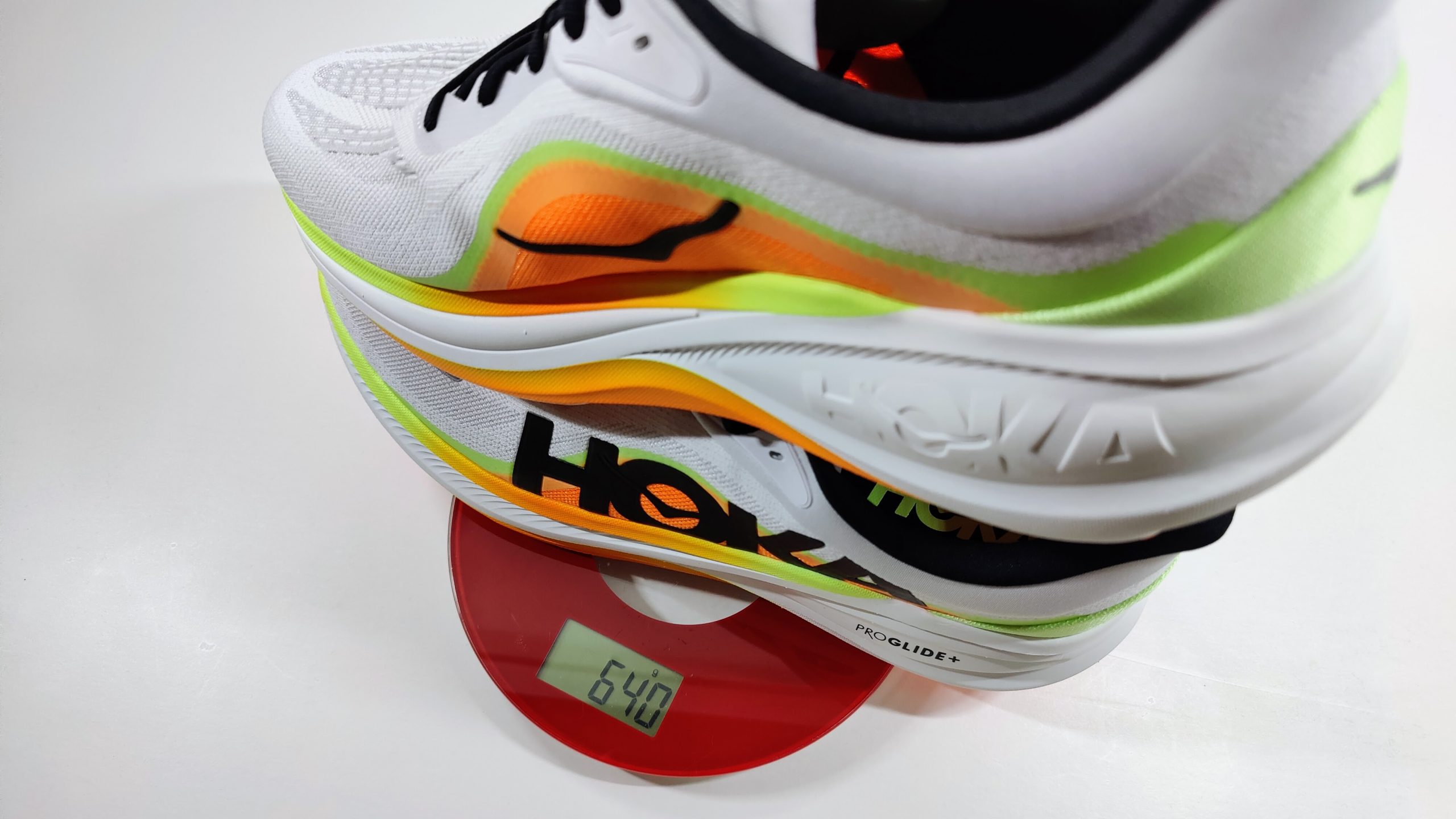 Two white running shoes with orange and lime green midsole on a red digital scale showing 640 grams, white background.