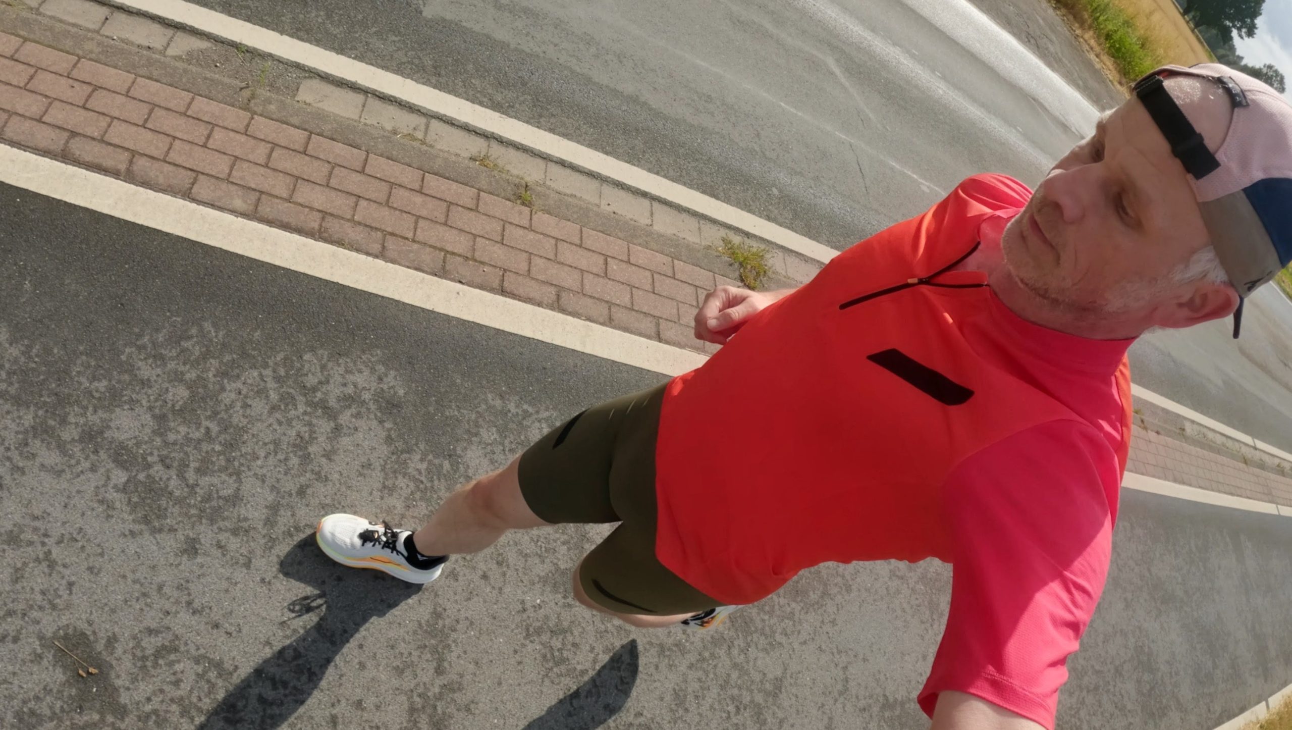 Runner in a red shirt and dark shorts photographed from a selfie perspective while running on asphalt next to a brick-paved bike path.