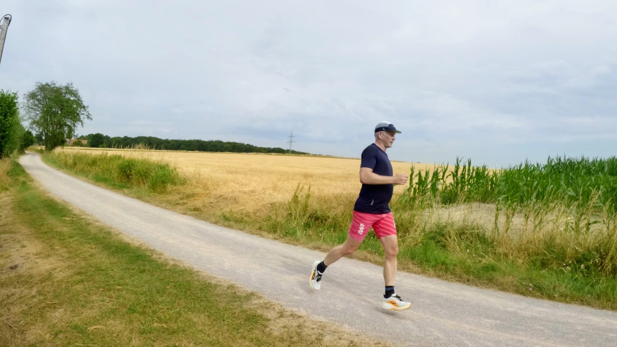 Runner in a dark shirt and pink shorts running on a gravel path between a harvested grain field and a corn field under a cloudy sky.