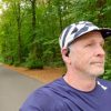 Man in a cap and navy running shirt with an open-ear headphone on a tree-lined path in autumn forest