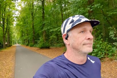Man in a cap and navy running shirt with an open-ear headphone on a tree-lined path in autumn forest