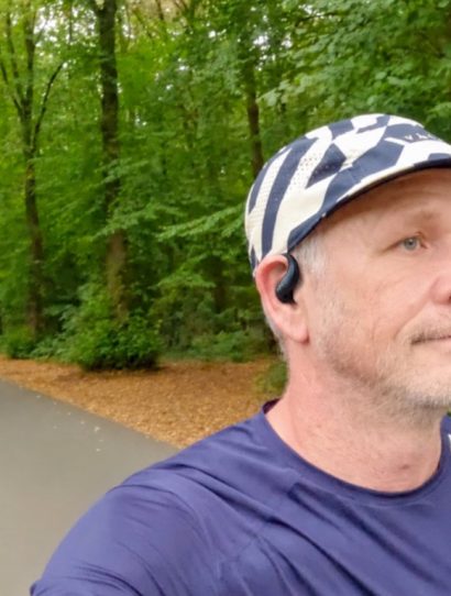Man in a cap and navy running shirt with an open-ear headphone on a tree-lined path in autumn forest
