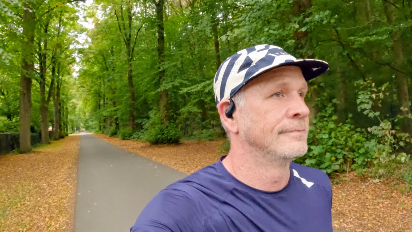 Man in a cap and navy running shirt with an open-ear headphone on a tree-lined path in autumn forest