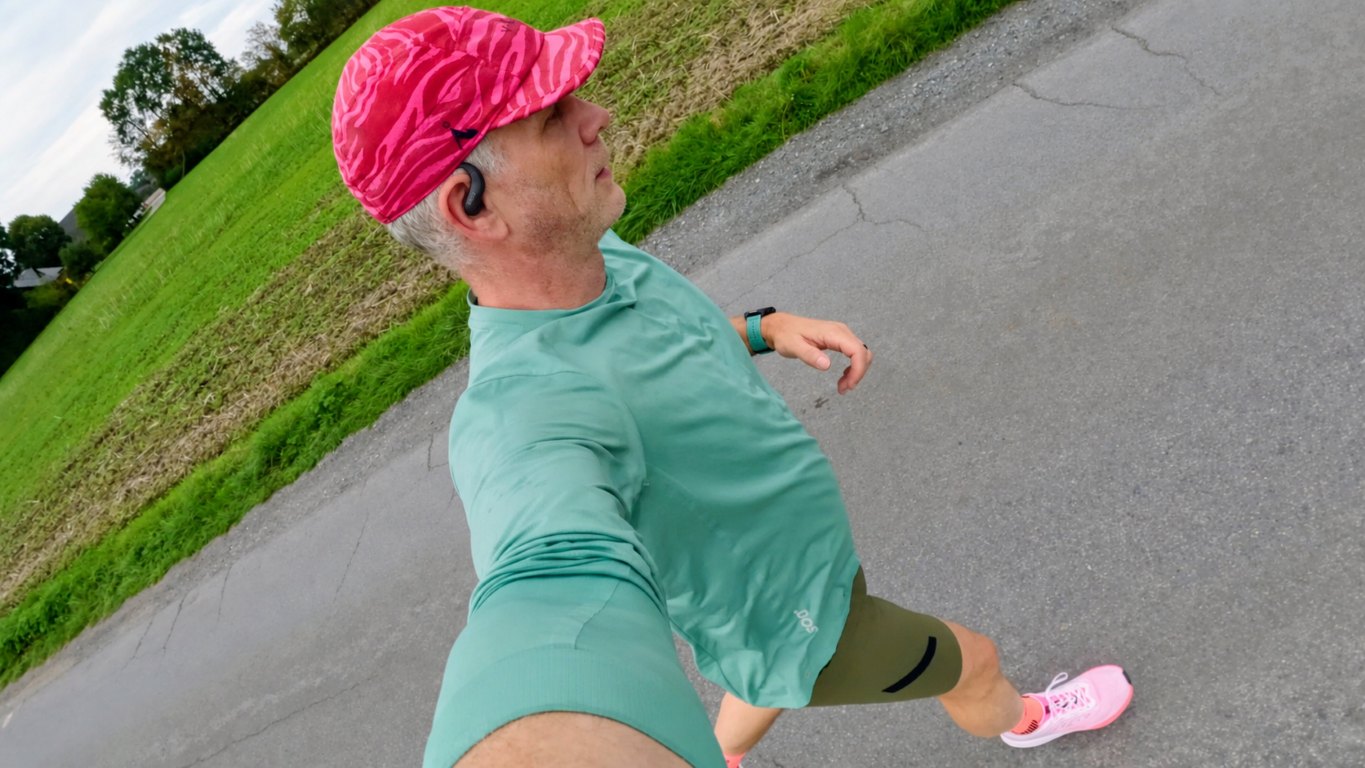 Man running on a country road in a green shirt and pink cap wearing an open-ear headphone, selfie perspective