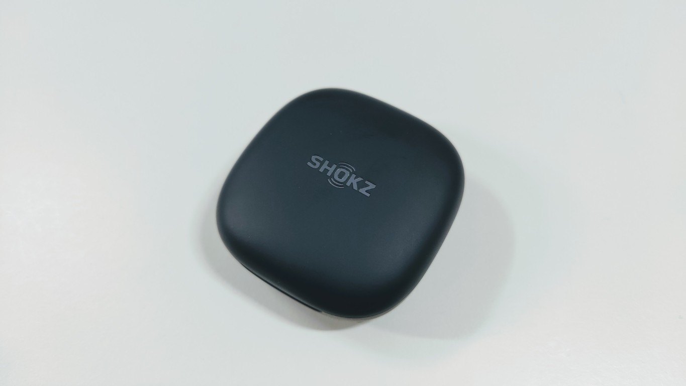 Dark grey charging case of the Shokz OpenFit Air 2 with logo on the lid, placed on a white surface
