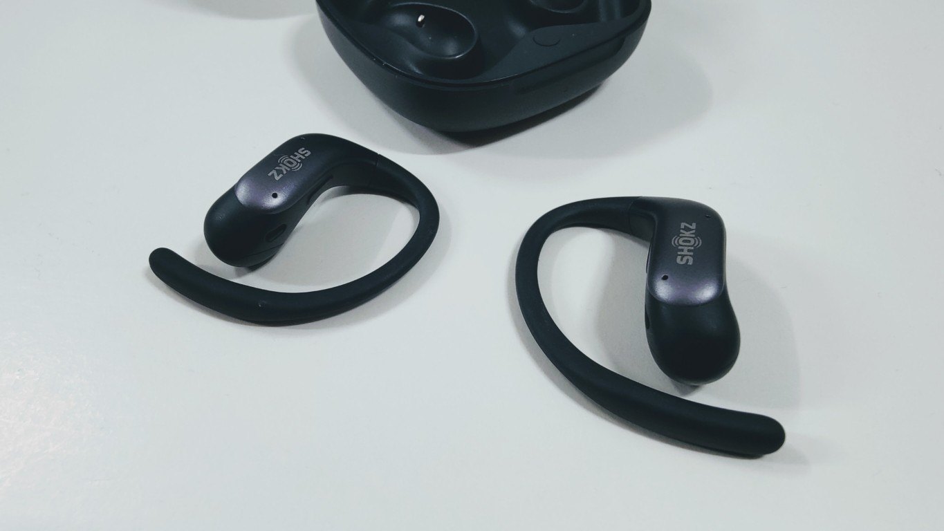 Two black open-ear earbuds with ear hooks lying next to the charging case on a white surface