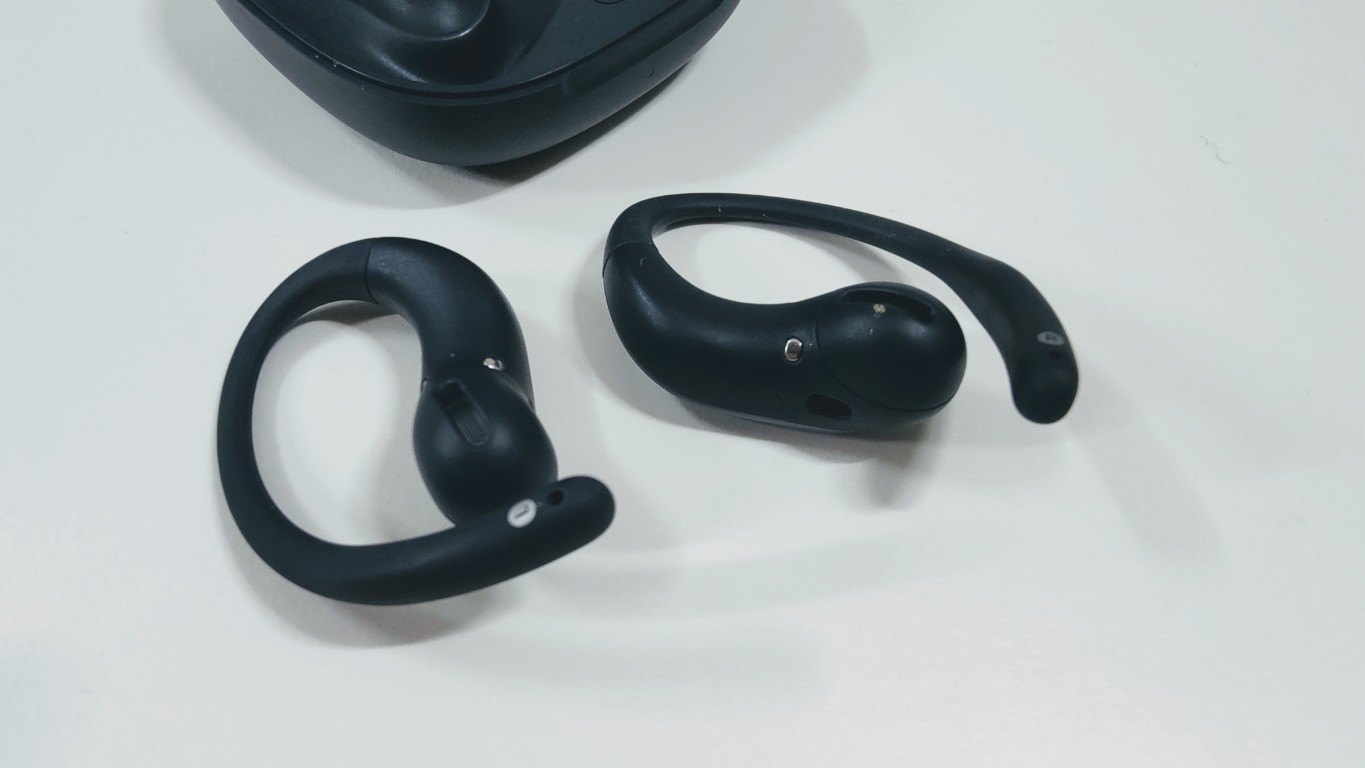 Black open-ear earbuds seen from behind, showing the inner side and contact pins, on a white surface